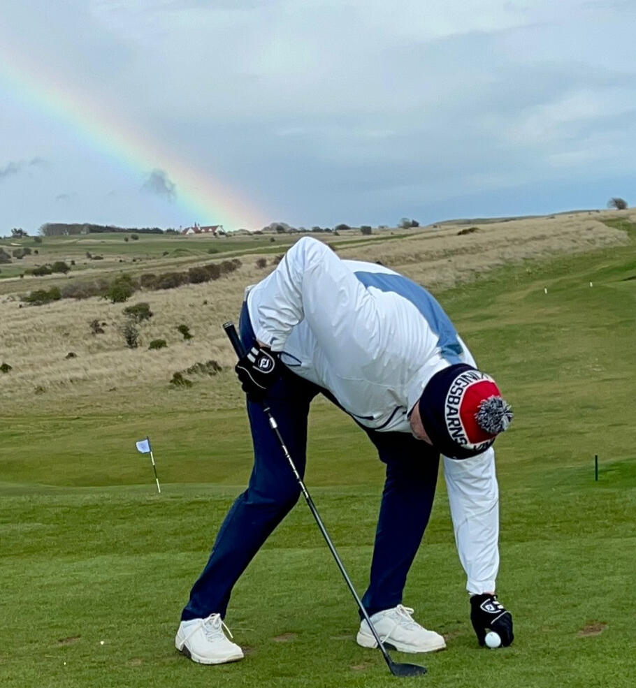 William bends to place a golf ball on a tee as a rainbow arcs across the course behind him.